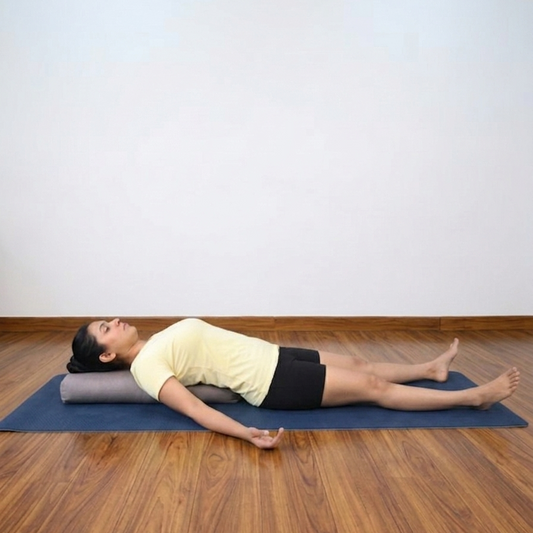 A woman lies on a blue yoga mat on a wooden floor, resting on a gray cylindrical pranayama pillow. She is wearing a pale yellow t-shirt and black shorts in a minimalist room with a white wall.