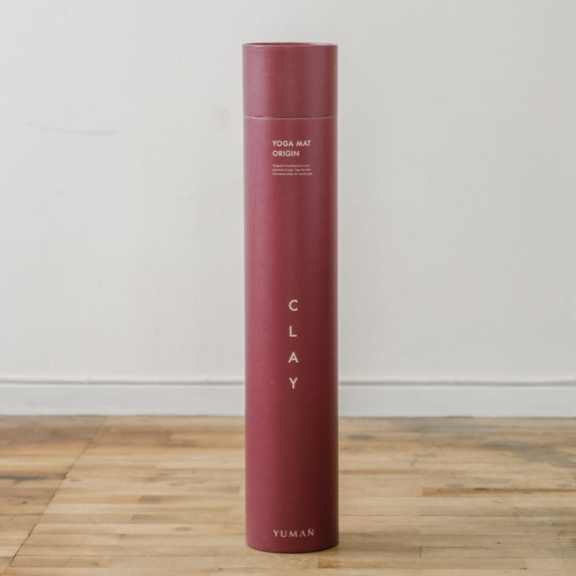 A tall, slim cylindrical yoga mat package or box in a muted clay pink color stands upright on a light wood floor against a plain off-white wall. The front has vertical text reading 'CLAY' and smaller text near the top that appears to say 'YOGA MAT ORIGIN' with 'YUMAN' at the bottom.