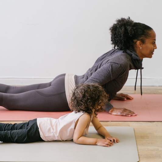 A woman and a young child are doing a yoga pose on mats in a bright studio with a white wall and wood floor.