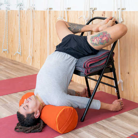 yogi lying on an orange yoga bolster with a yoga chair behind them on a red mat.