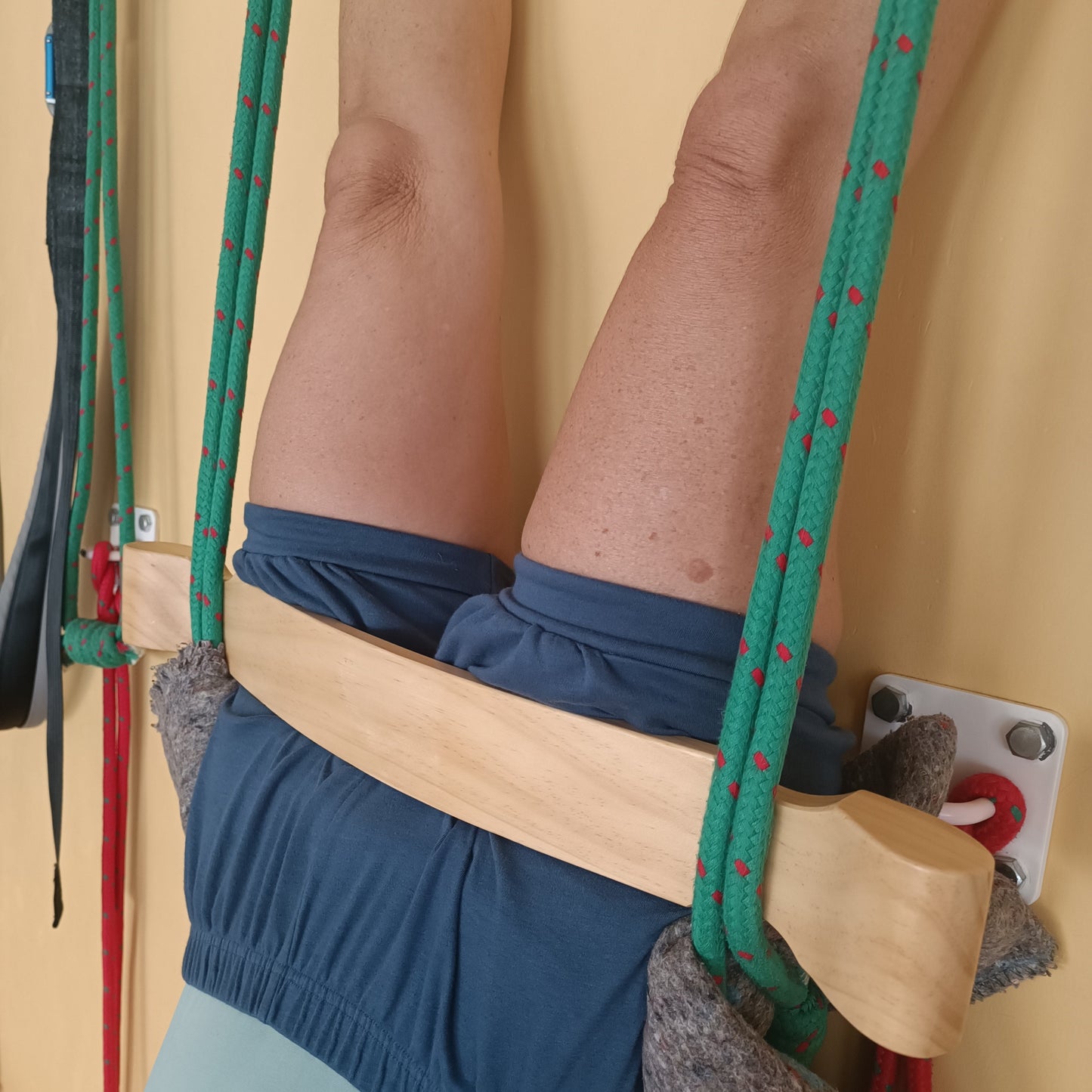 A close-up of a person using a wall-mounted iyengar yoga ropes and a curvature plank, with both legs extended upward against a beige wall. The person is wearing blue yoga shorts.