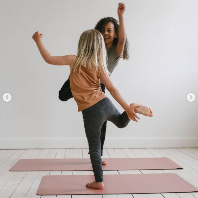 Two girls in athletic wear pose together on pink yoga mats in a bright, minimalist room with white walls and light wood floors.