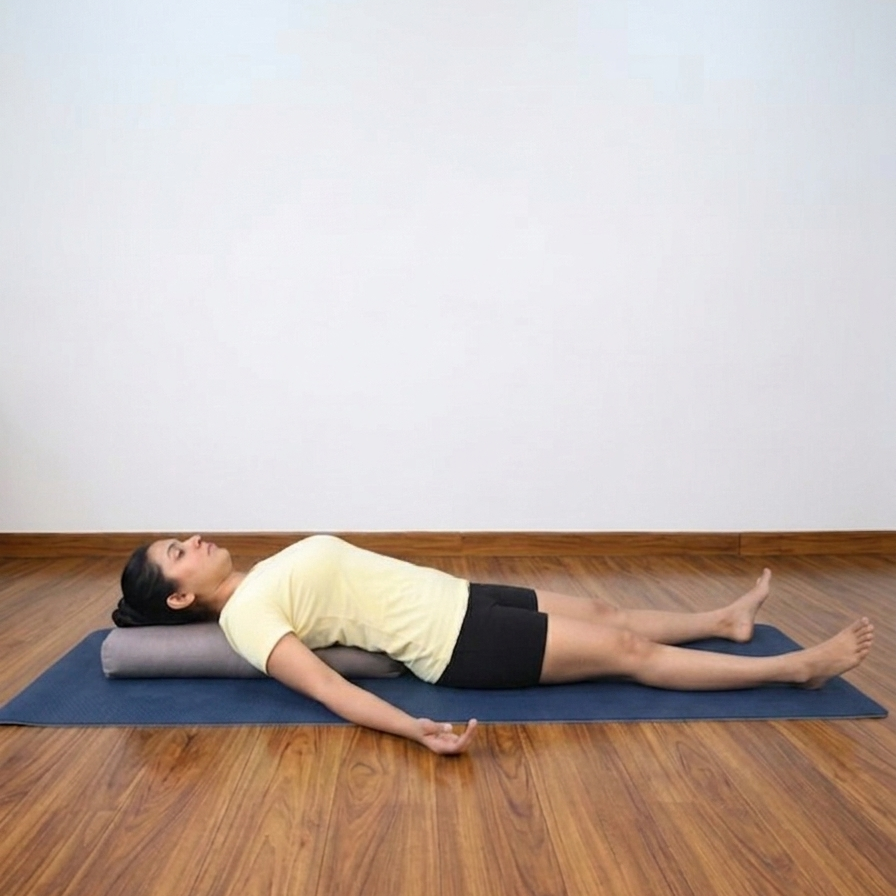 A woman lies on a blue yoga mat on a wooden floor, resting on a gray cylindrical pranayama pillow. She is wearing a pale yellow t-shirt and black shorts in a minimalist room with a white wall.