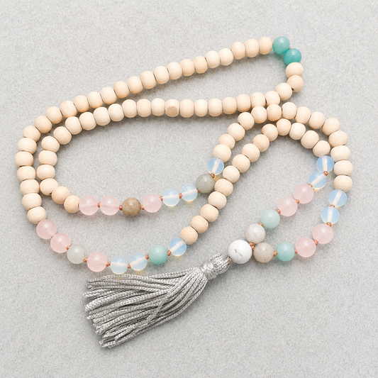 A japa mala with light wood beads, rose quartz and mint green amazonite stones, iridescent white beads, and a silver tassel, displayed on a neutral gray background.