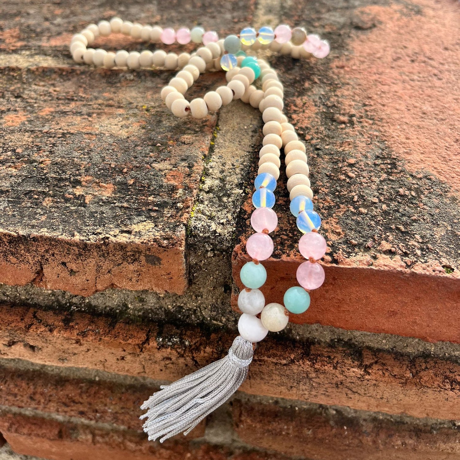 A japa mala with a gray tassel draped over a rustic brick ledge, featuring cream-colored wooden beads, pastel pink, blue, and mint green glass beads.