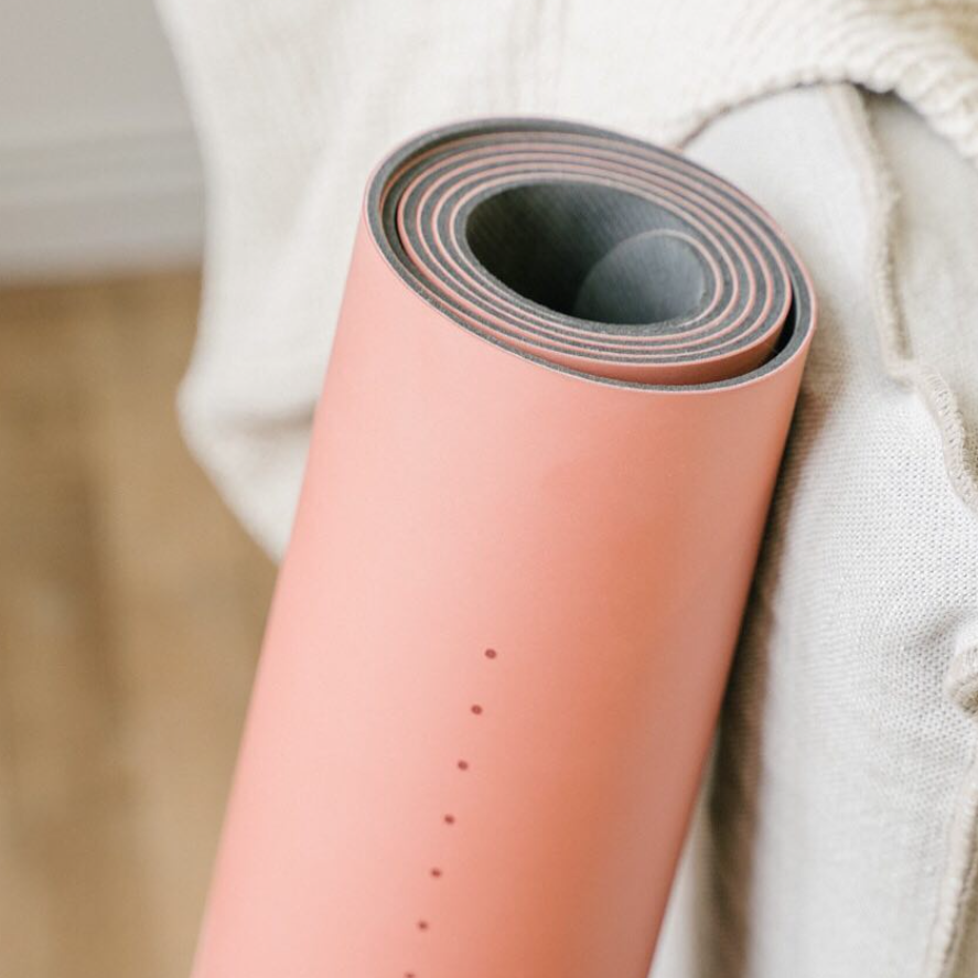 A rolled pink yoga mat with a dark gray underside, resting against a textured cream linen throw blanket on a wooden floor.
