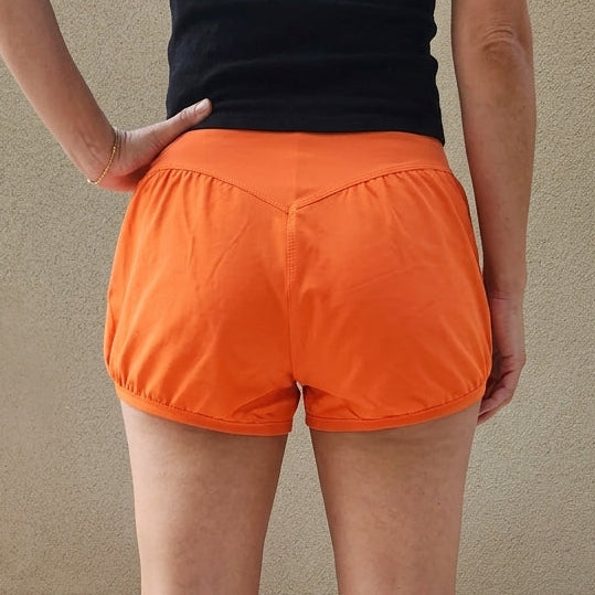 A woman viewed from behind wearing bright orange bloomer yoga shorts and a black short-sleeve top, standing barefoot against a beige wall.
