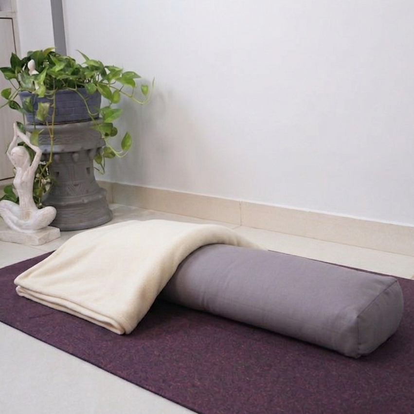A gray cylindrical pranayama bolster pillow resting on a dark purple yoga mat, with a folded cream blanket 