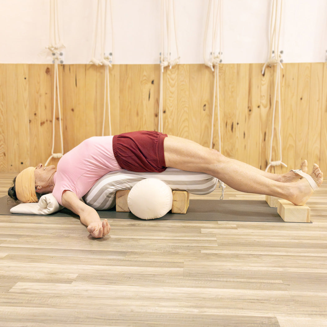 Person lying on a yoga bolster wearing burgundy yoga shorts in a wooden room with iyengar yoga props.