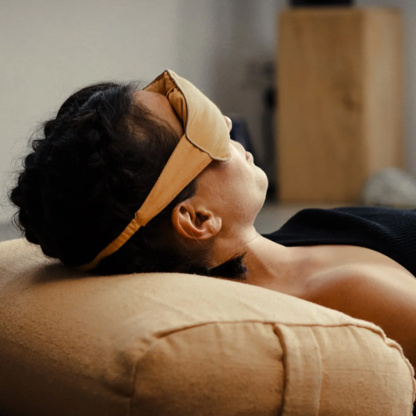 A person lying down with their head resting on a beige pillow, wearing a yellow sleep mask. The background is softly blurred, showing a neutral indoor setting.